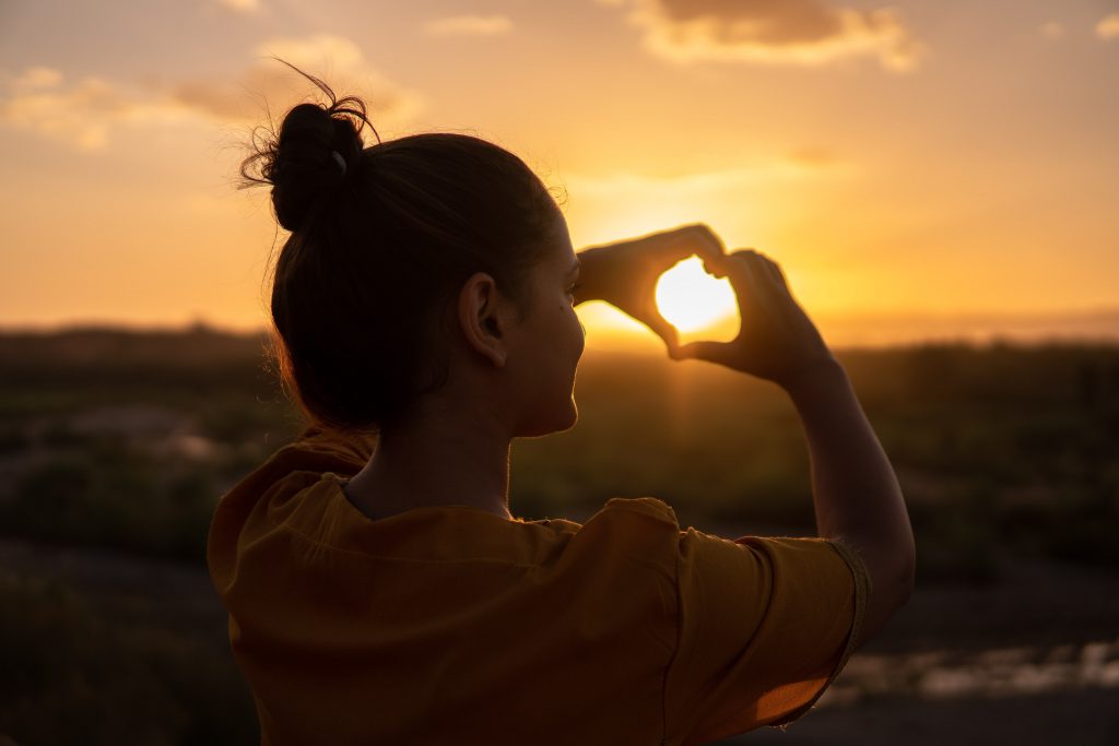 Seorang Wanita, Memandang Ke Arah Matahari saat Senja Sambil Tangannya Membentuk Simbol Love